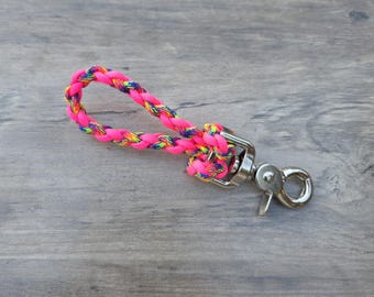 May include: A bright pink braided keychain wrist strap with a rainbow pattern woven throughout. The strap is attached to a silver-colored metal clasp. The background is a gray wooden surface.