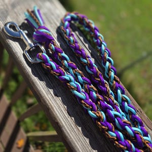 May include: A braided dog leash made of purple, light blue, and brown patterned paracord. The leash features a silver-colored metal swivel snap hook at one end and a braided loop handle at the other, showcasing a thick, multi-strand weave design.