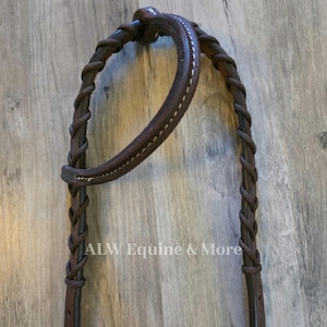 May include: Close-up of a brown leather horse bridle with white stitching and braided accents. The bridle features a curved browband and cheek pieces. The text "ALW Equine & More" is visible.
