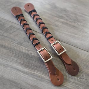 Op de afbeelding: Twee bruine leren riemen met een zwart koord dat in een kruispatroon door voorgesneden gaten is gevlochten. Elke riem bevat een zilverkleurige metalen gesp voor verstelling en een afgerond uiteinde met een enkel gat voor bevestiging.
