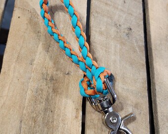 May include: A wrist lanyard crafted from braided turquoise and orange paracord, featuring a silver-toned metal swivel lobster clasp at the base. The cord is woven into a continuous loop, securely knotted to the metal hardware at the bottom end.
