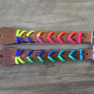 Op de afbeelding: Twee bruine leren riemen met een gevlochten ontwerp van neon paracord. De bovenste riem heeft gele, paarse, oranje en roze koorden, terwijl de onderste riem gele, paarse, groene en blauwe koorden heeft. Beide hebben zilverkleurige metalen gespen.