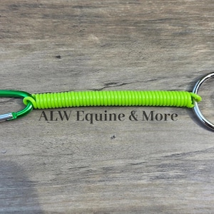 May include: A bright green coiled lanyard with a green carabiner clip and a silver split ring. The text "ALW Equine & More" is printed on the wooden surface.