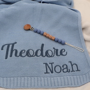 May include: A light blue knitted baby blanket with the embroidered names "Theodore Noah". A wooden and blue pacifier is resting on the blanket. The blanket is folded and has a ribbed border.