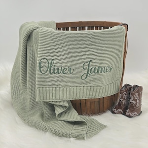 May include: A sage green knitted baby blanket with the embroidered name "Oliver James" in a script font. The blanket is draped over a wooden basket, with a pair of brown baby cowboy boots beside it. The blanket is made of a soft, textured material.