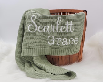 May include: A sage green knitted baby blanket with the name "Scarlett Grace" embroidered in white. The blanket is folded over a brown wicker basket, with a portion of the blanket draped over the side. The blanket is made of a soft, textured material.