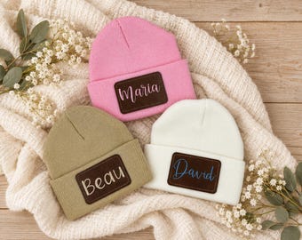 May include: Three knit beanies in pink, olive green, and white, each featuring a rectangular dark brown leather patch. The pink beanie displays the name Maria, the olive green beanie shows Beau, and the white beanie features the name David.