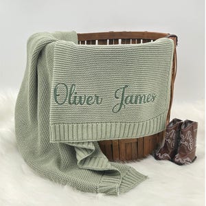 May include: A sage green knitted baby blanket with the embroidered name "Oliver James" is draped over a wooden basket. Two small brown cowboy boots sit beside the basket on a white, fluffy surface.