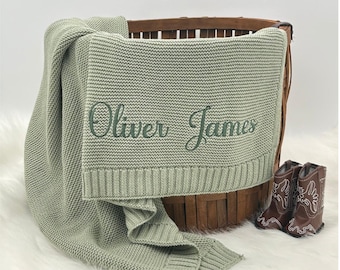 May include: A sage green knitted baby blanket with the embroidered name "Oliver James" is draped over a wooden basket. Two small brown cowboy boots sit beside the basket on a white, fluffy surface.