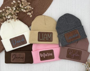 May include: A collection of six knit beanies in white, tan, dark gray, dark brown, pink, and mauve. Each beanie features a rectangular brown leather patch engraved with a name: David, Liam, Lincoln, James, Maria, and Rylee, respectively.