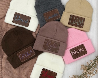 May include: A collection of seven knit beanies in white, dark gray, tan, dark brown, mauve, pink, and white. Each beanie features a brown rectangular leather patch with a name engraved in various colors: David, Lincoln, Liam, Grace, Ryleo, Maria, and Hayes.
