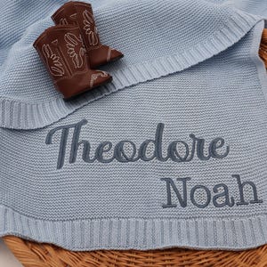 May include: A light blue knitted baby blanket with the names "Theodore" and "Noah" embroidered in gray thread.