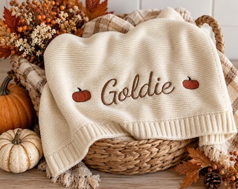 May include: A cream-colored knit blanket featuring the embroidered name Goldie in brown script, flanked by two small orange pumpkins. The blanket is draped over a woven basket containing autumn foliage and a plaid fabric liner.