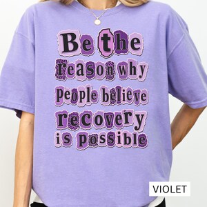 以下が含まれることがあります： 「Be the reason why people believe recovery is possible」という黒い文字がプリントされたバイオレットカラーの半袖Tシャツ。各単語は、破れた紙のような縁取りが施された、紫とヒョウ柄の長方形の中に配置されています。