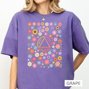 以下が含まれることがあります： グレープパープルのTシャツ。中央に「Unity, Service, Recovery」の文字が入ったオレンジ色の三角形のエンブレムが配置されています。その周囲は、ピンク、黄色、青、白の色合いの様々な小さな花々が密集したカラフルなパターンで飾られています。
