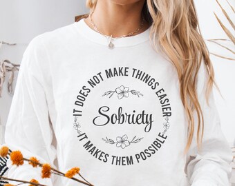 May include: White long-sleeved t-shirt with a black circle graphic that says "It does not make things easier, It makes them possible" with the word "Sobriety" in a larger font inside the circle.
