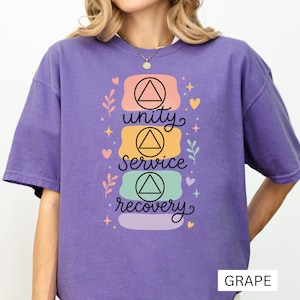 以下が含まれることがあります： 紫色のTシャツで、黒い三角形のシンボルが入った3つのパステルカラーの長方形が重なったグラフィックが特徴です。筆記体で「unity」「service」「recovery」と書かれており、小さなハート、星、葉のイラストが周囲に描かれています。