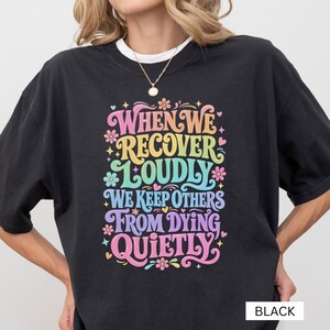 以下が含まれることがあります： 「WHEN WE RECOVER LOUDLY WE KEEP OTHERS FROM DYING QUIETLY」というテキストがスタイリッシュなフォントでプリントされた黒のTシャツ。ピンク、黄色、青の色合いの小さな花や星のアイコンが周囲にデザインされています。