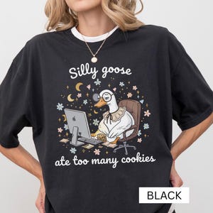 Könnte beinhalten: Schwarzes T-Shirt mit einer skurrilen Grafik einer Gans mit Brille und Rüschenkragen, die an einem Schreibtisch mit Computer und Keksen sitzt. Der Text lautet "Silly goose ate too many cookies."