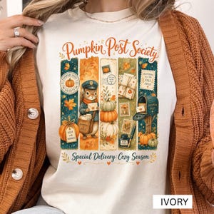 May include: An ivory t-shirt featuring a graphic print with the text Pumpkin Post Society and Special Delivery: Cozy Season. The design includes a squirrel mail carrier, pumpkins, envelopes, a mailbox, stamps, and an inkwell in autumn colors.