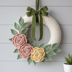 May include: A cream-colored crochet wreath adorned with pink and yellow crochet roses and green leaves. A green velvet ribbon is tied in a bow at the top. The wreath is hanging on a light gray wall.