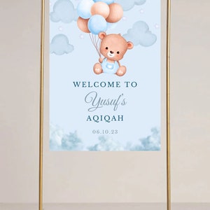 May include: A blue and white welcome sign with a teddy bear holding balloons. The sign says "Welcome to Yusuf's Aqiqah 06.10.23".