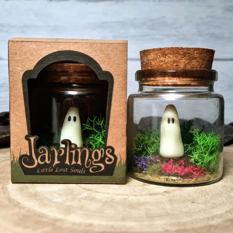 May include: A small glass jar with a cork lid contains a white ghost figurine standing in a bed of green moss. The jar is labeled "Jarlings Little Lost Souls".