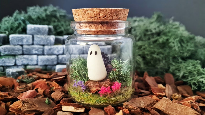 May include: A small glass jar with a cork lid contains a miniature white ghost figurine standing in a bed of green moss and pink flowers.