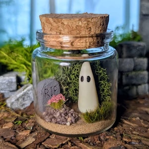 May include: A small glass jar with a cork stopper, containing a miniature Halloween scene. Inside, a white ghost figure stands near a grey tombstone with "RIP" inscribed on it. The scene includes sand, moss, and small pink flowers.