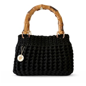 May include: A black, hand-knitted handbag with a bamboo handle. The bag features a chunky knit design and gold-colored metal hardware. A small, round charm with a heart design hangs from the side.