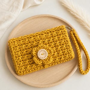May include: A mustard yellow crochet wristlet pouch featuring a textured stitch pattern and a matching crocheted flower embellishment at the center. A round wooden button is sewn onto the flower, inscribed with the text Handmade with love.