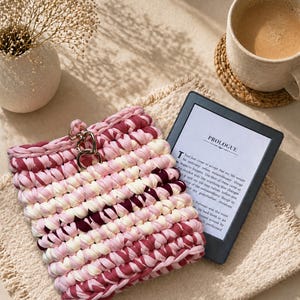 May include: A handmade, pink and white crocheted case with a silver clasp, next to an e-reader displaying the text "PROLOGUE". A cup of coffee sits on a woven coaster, with a vase of dried flowers in the background.