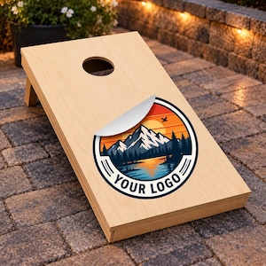 May include: A light-colored wooden cornhole board featuring a circular graphic with a mountain landscape, a sunset, and a lake. The graphic includes the text YOUR LOGO at the bottom. A partially peeled white sticker is visible on the board surface.