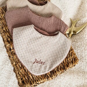 Puede incluir: Un conjunto de tres baberos triangulares para bebé apilados. El babero superior presenta un patrón de cuadros vichy en beige y blanco con el nombre Jules bordado en hilo marrón. El babero central es de color rosa empolvado y el inferior tiene rayas beige.