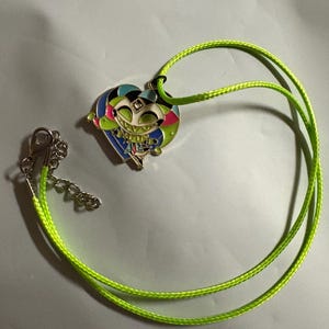 May include: A heart-shaped pendant necklace with a cartoon character design. The pendant features a smiling face with colorful accents and is suspended from a neon green cord necklace with a silver clasp.