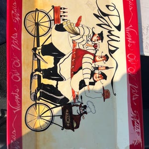 Puede incluir: Una bandeja de servicio rectangular de cerámica con un centro color crema y un borde rojo ancho. El borde presenta el texto repetido Paris, Vivants y Oui Oui. El centro representa a tres figuras en una bicicleta tándem con vino y pan.