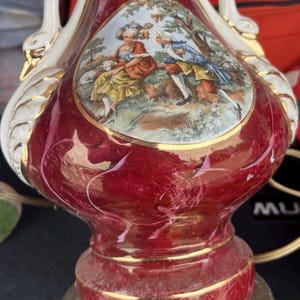 Puede incluir: Lámpara de cerámica roja vintage con base dorada y asas en forma de cisne. La lámpara presenta una escena pintada de personas en un jardín. La lámpara tiene un acabado brillante y es un artículo de decoración del hogar.