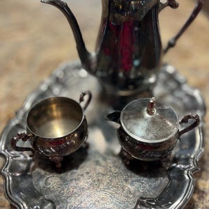 Może przedstawiać: Srebrny zestaw do herbaty na ozdobnej tacy. Zestaw zawiera czajniczek, cukiernicę z pokrywką i małą śmietankę. Taca i zestaw mają bogate zdobienia.