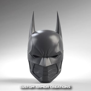 Op de afbeelding: Een donkergrijs Batman-masker met puntige oren en een gedetailleerd, hoekig ontwerp. Het masker heeft een getextureerd oppervlak en bedekt het hele gezicht. De tekst "CUSTOM ARMOR CREATIONS" staat onderaan.