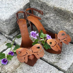 May include: Rusty metal high-heeled shoe sculptures with butterfly details, decorated with purple flowers and green leaves. The shoes feature ankle straps and are presented on a grey stone surface, creating a rustic, artistic display.