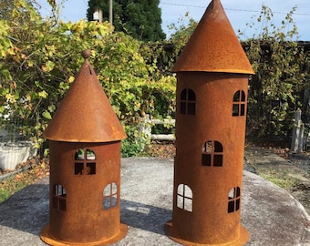 Könnte beinhalten: Zwei rostige Metallturm-Skulpturen mit spitzen Dächern und ausgeschnittenen Fenstern. Die Türme sind unterschiedlich groß und stehen auf einer Steinoberfläche.