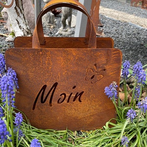 Könnte beinhalten: Ein rostiger Metall-Pflanzkasten in Form einer Einkaufstasche mit einem ausgeschnittenen Schmetterlingsmotiv und dem Wort "Moin" auf der Vorderseite. Der Pflanzkasten steht auf einem Bett aus grünem Gras und lila Blumen.