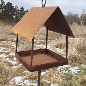 May include: A rusty metal bird feeder with a square tray and a peaked roof. The feeder is mounted on a metal pole and is standing in a field of brown grass and snow.