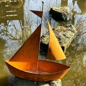 Op de afbeelding: Een roestige metalen zeilboot-sculptuur met een puntige boeg en een enkel zeil. De sculptuur staat op een rots in een vijver.
