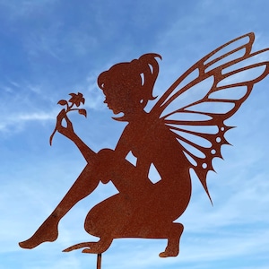 Op de afbeelding: Een roestig metalen fee-figuur met vleugels, die een bloem vasthoudt. De fee zit op een metalen standaard en is gesilhouetteerd tegen een blauwe lucht.