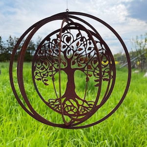Op de afbeelding: Een roestbruine metalen levensboomsculptuur met een cirkelvormig ontwerp. De boom is gecentreerd in een bol, omringd door twee extra concentrische ringen. De sculptuur hangt tegen een achtergrond van groen gras en een bewolkte lucht.