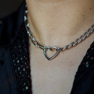 May include: A silver-toned metal chain necklace featuring a central heart-shaped link. The necklace is constructed from interlocking oval links that connect to the open heart charm, creating a continuous metallic loop around the neck area.