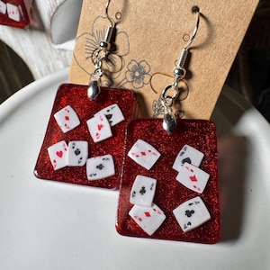Puede incluir: Un par de pendientes rectangulares con una base de resina roja brillante incrustada con naipes blancos en miniatura. Cada pendiente cuelga de un gancho de metal plateado y muestra varios palos de cartas, incluyendo corazones, diamantes y tréboles.