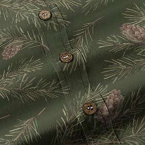 May include: Green button-up shirt with a pine needle and pine cone pattern. The shirt features wooden buttons and a detailed nature-inspired design. The fabric has a slightly textured appearance.