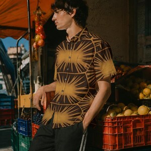 May include: A short-sleeved button-down shirt featuring a repeating geometric pattern of dark brown and golden yellow sunburst motifs. The shirt has a standard collar, a front button placket, and a single chest pocket on the left side.